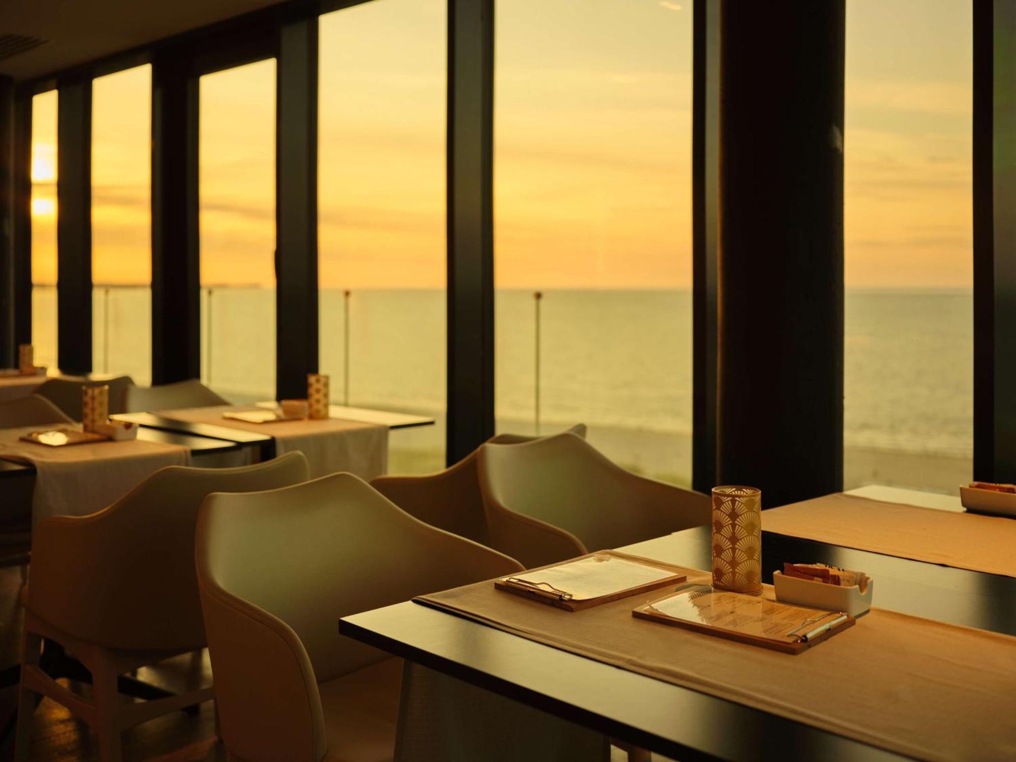 Radisson Blu Resort Świnoujście מראה חיצוני תמונה The photo shows a restaurant interior with a beautiful view of the ocean during sunset. The large windows allow natural light to flood the space, highlighting the warm colors of the sunset in the sky. There are several tables, each set with placemats