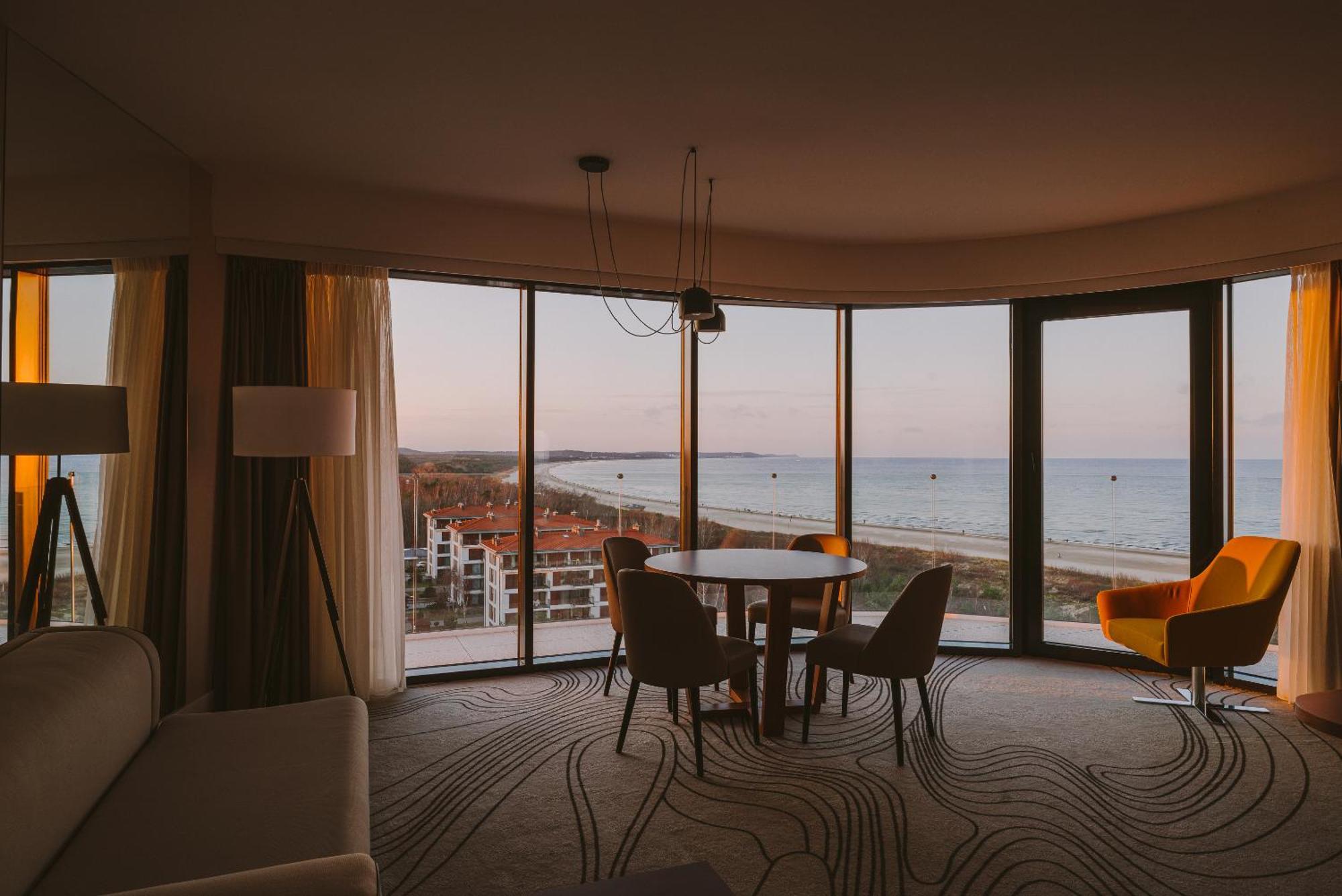Radisson Blu Resort Świnoujście מראה חיצוני תמונה The photo shows a modern and stylish interior of a room featuring large windows that provide a view of the beach and ocean. The room is furnished with a round table and chairs, along with a cozy sofa and a bright yellow armchair. The decor has a cont