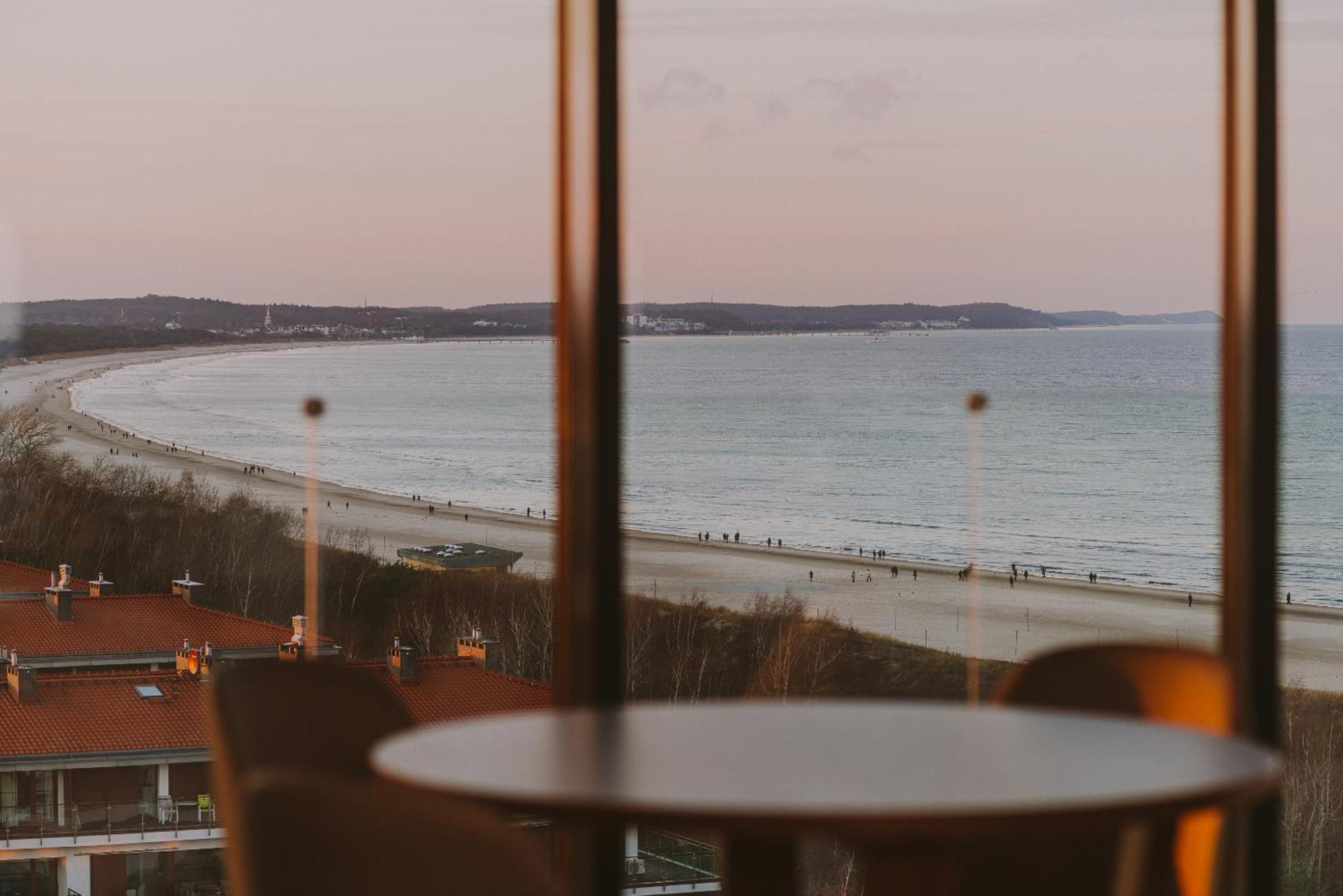 Radisson Blu Resort Świnoujście מראה חיצוני תמונה The photo shows a picturesque view of a beach scene from indoors. In the foreground, there is a round table with some chairs around it, suggesting a cozy interior space. Through a large window, you can see the beach with gentle waves hitting the shor