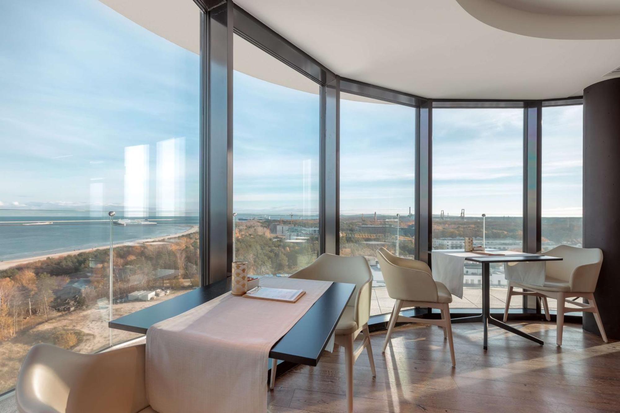 Radisson Blu Resort Świnoujście מראה חיצוני תמונה The photo shows a modern dining area with large windows that offer a panoramic view of the landscape outside, including a body of water and possibly a shoreline. The design features sleek furniture, including a table with chairs arranged around it, a