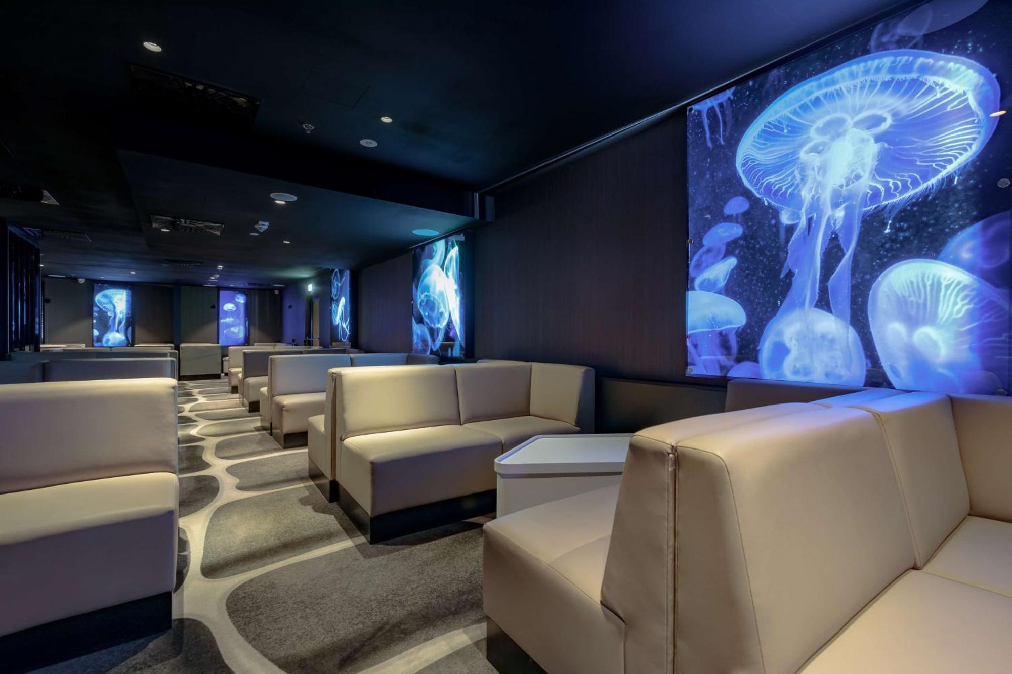 Radisson Blu Resort Świnoujście מראה חיצוני תמונה The photo shows a modern lounge space with a sleek design. The area features light-colored, upholstered seating arranged in an open layout. The walls are decorated with large, vibrant images of jellyfish illuminated in blue, creating a calming underw