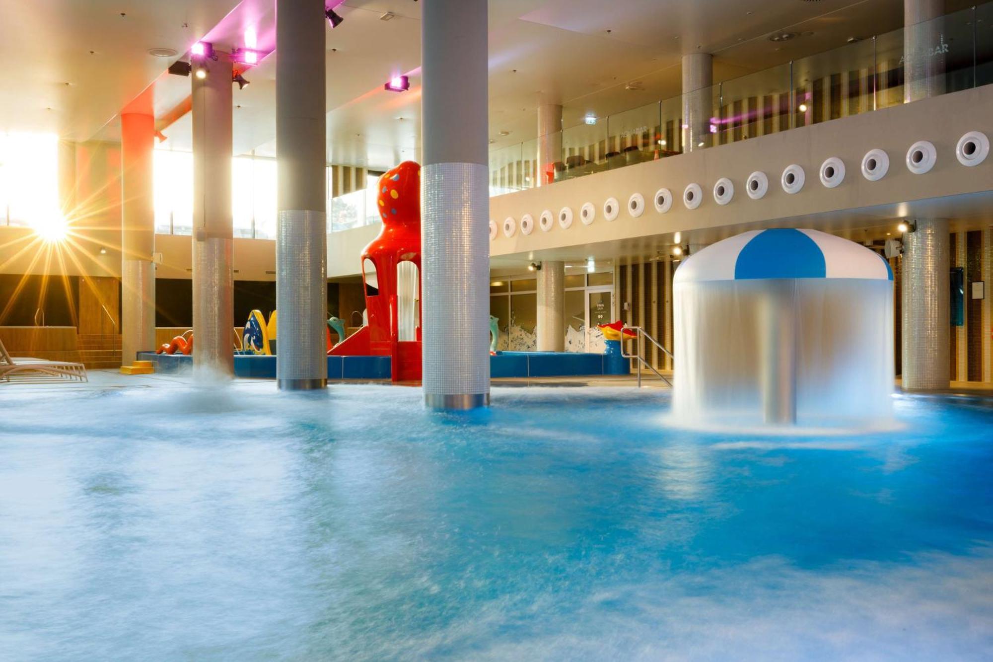 Radisson Blu Resort Świnoujście מראה חיצוני תמונה The photo shows an indoor water park or pool area. There is a large body of water with playful features, including a water slide shaped like a red bear and a smaller structure with a blue and white canopy. The lighting is bright, suggesting a sunny a