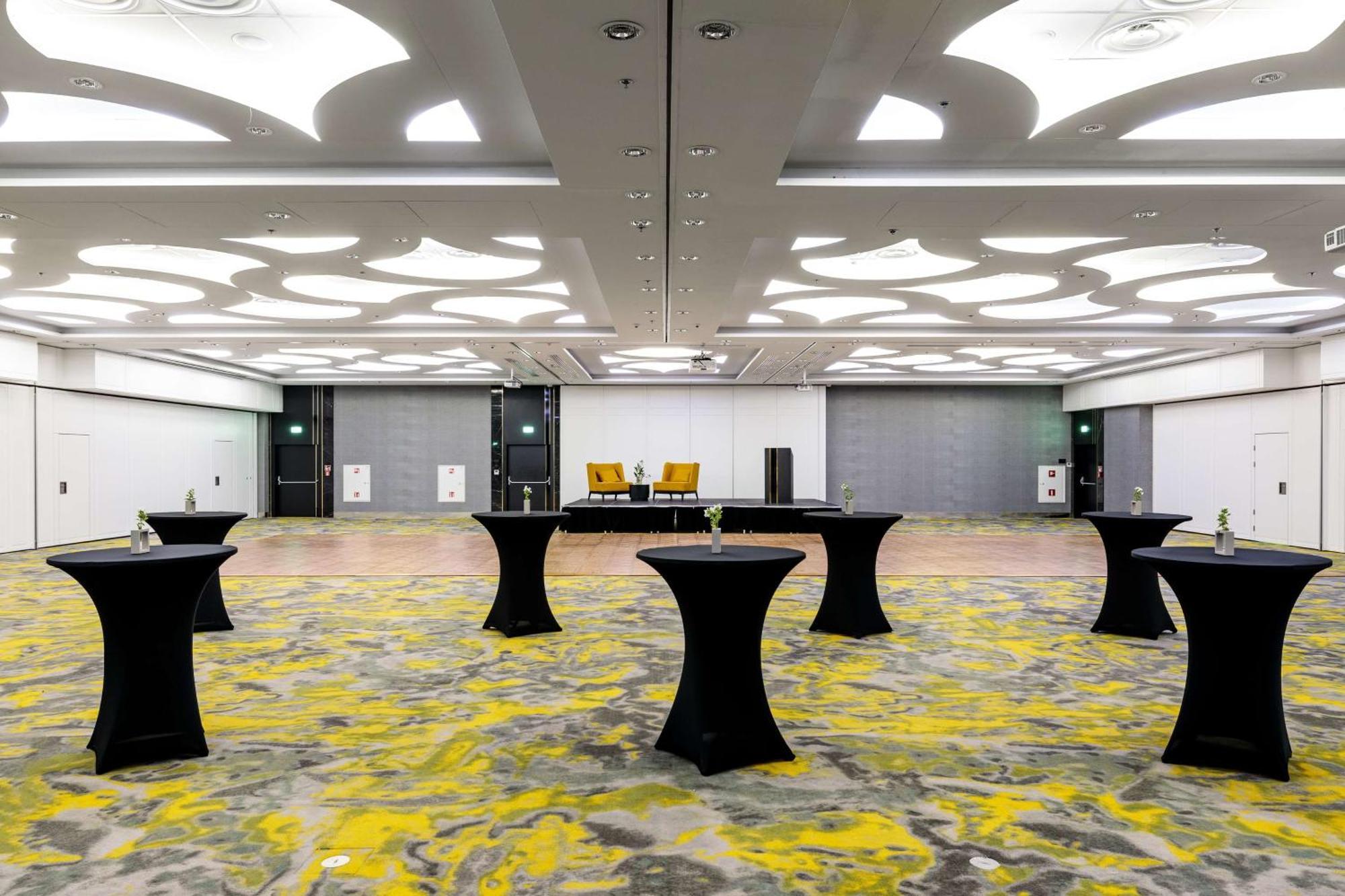 Radisson Blu Resort Świnoujście מראה חיצוני תמונה The photo shows a spacious interior room set up for an event. The flooring features a patterned design in gray with yellow accents. There are several black tall tables scattered throughout the area, and in the background, there are two large yellow a