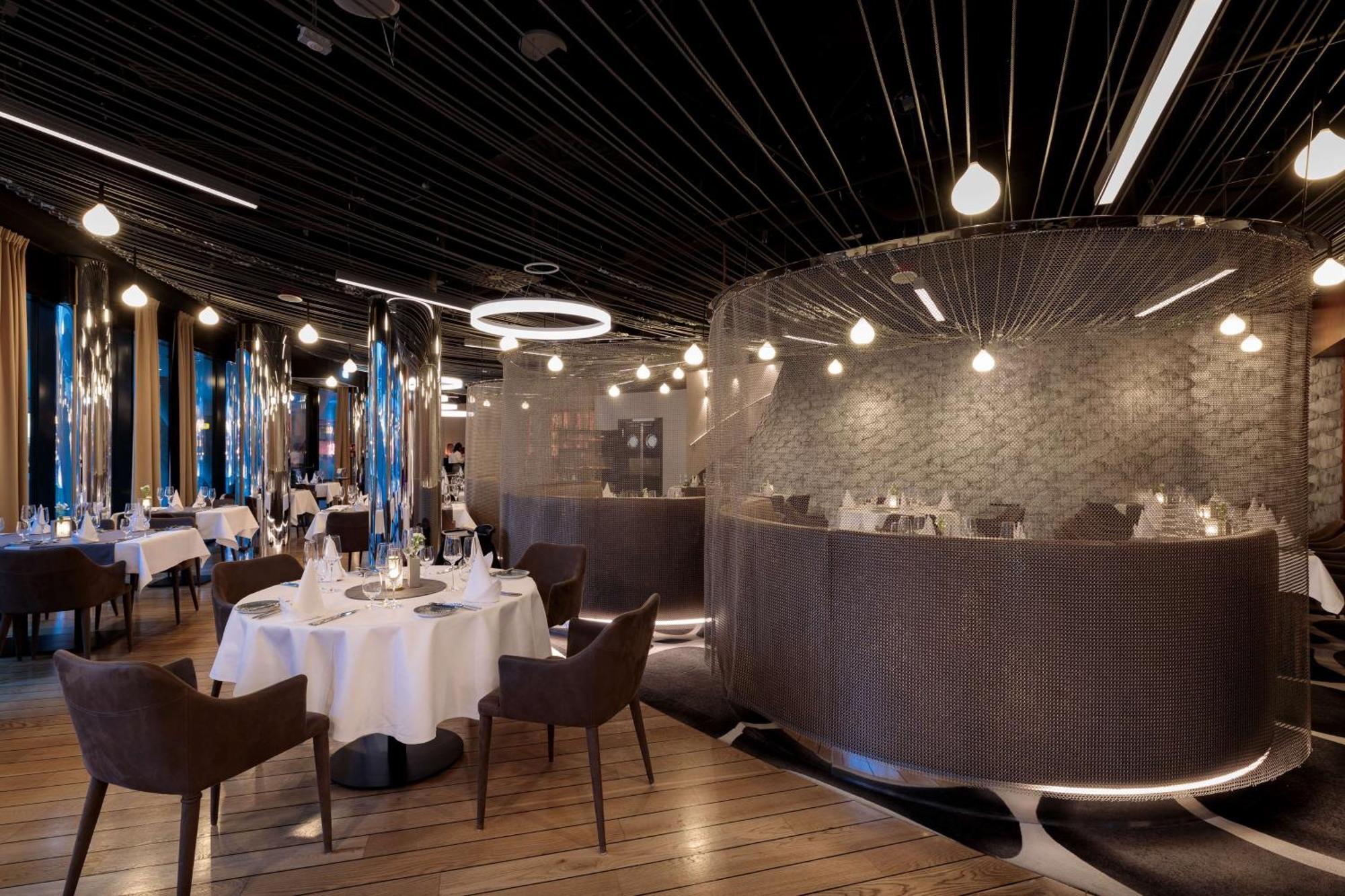 Radisson Blu Resort Świnoujście מראה חיצוני תמונה The photo shows an elegant restaurant interior. It features polished wooden floors and contemporary decor. Round tables are set with white tablecloths and have chairs arranged around them. The lighting is modern, with multiple pendant lights hanging 
