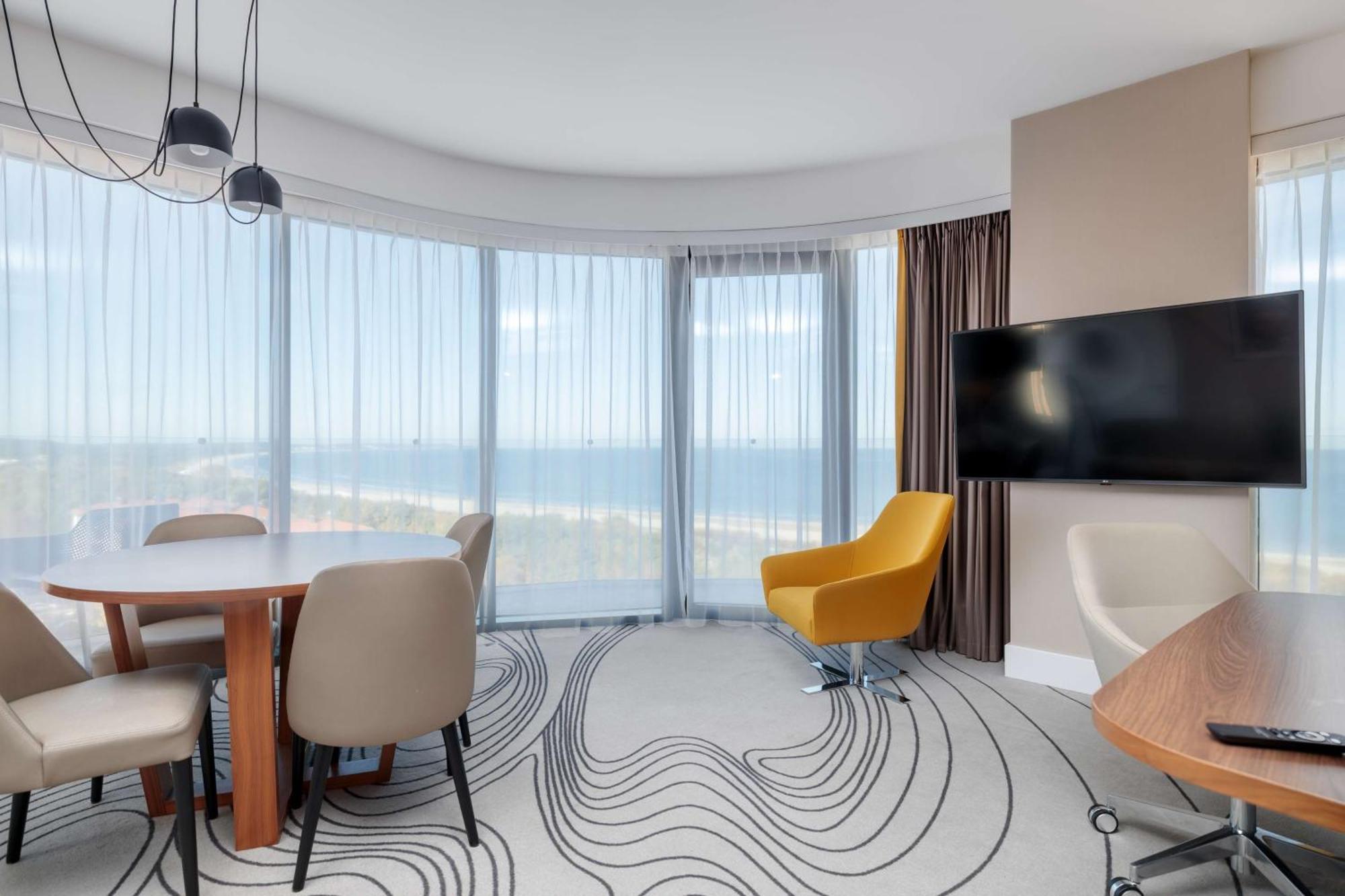 Radisson Blu Resort Świnoujście מראה חיצוני תמונה The photo shows a modern, stylish interior of a room with large windows that provide a panoramic view. The space features a round dining table surrounded by chairs, including a distinct yellow armchair. There is a flat-screen television mounted on th