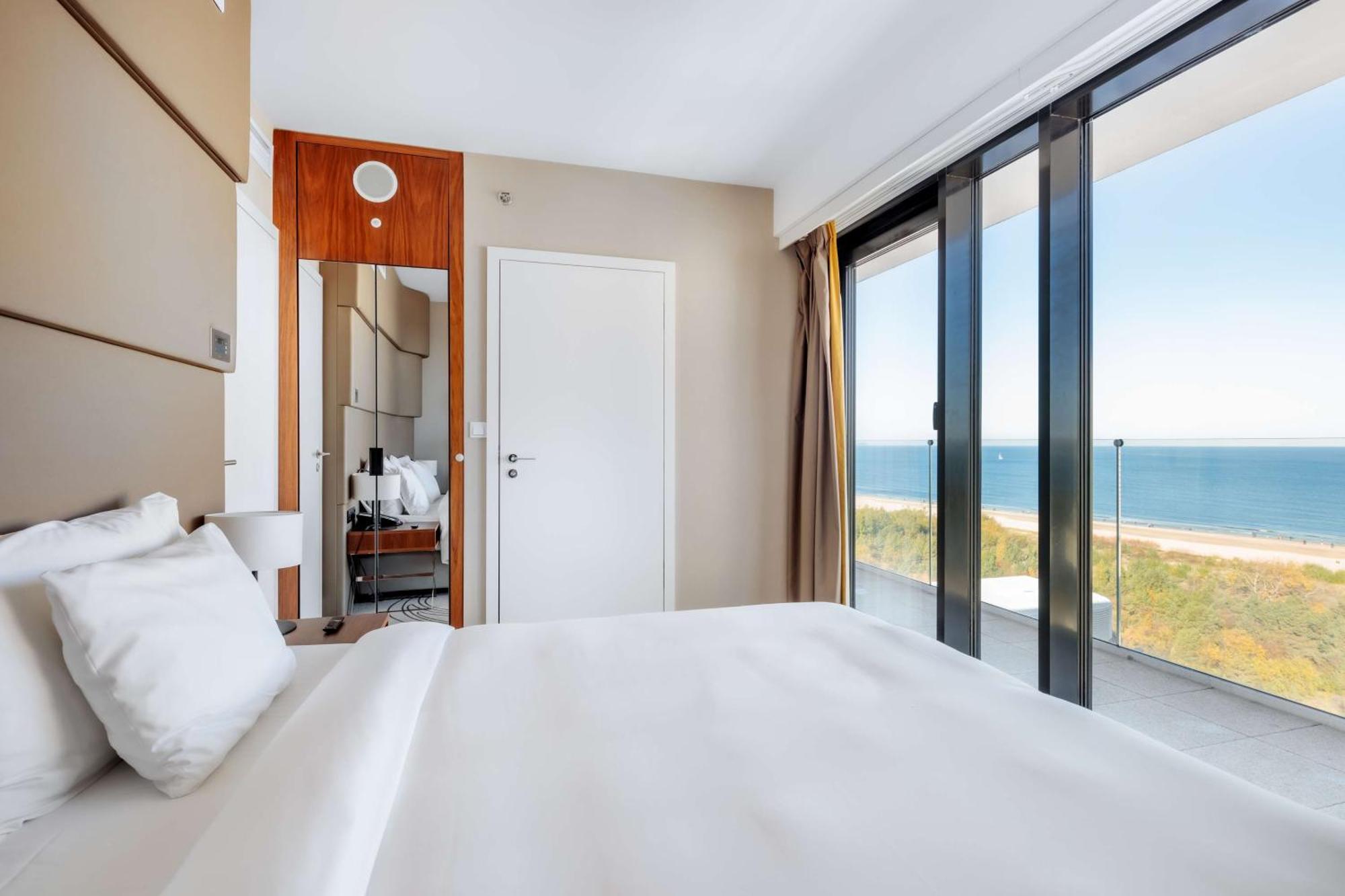 Radisson Blu Resort Świnoujście מראה חיצוני תמונה The photo shows a modern hotel room with a bright and airy atmosphere. There is a large bed with white bedding positioned near a glass wall that offers a view of the ocean. The room features neutral-colored walls and wooden accents, enhancing its con