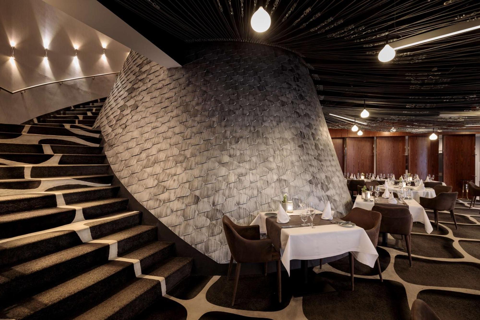 Radisson Blu Resort Świnoujście מראה חיצוני תמונה The photo shows the interior of a stylish restaurant. In the foreground, there are elegantly set tables with white tablecloths and dark chairs. A unique, curving stone wall adds a distinct architectural feature, and a grand staircase with a textured 