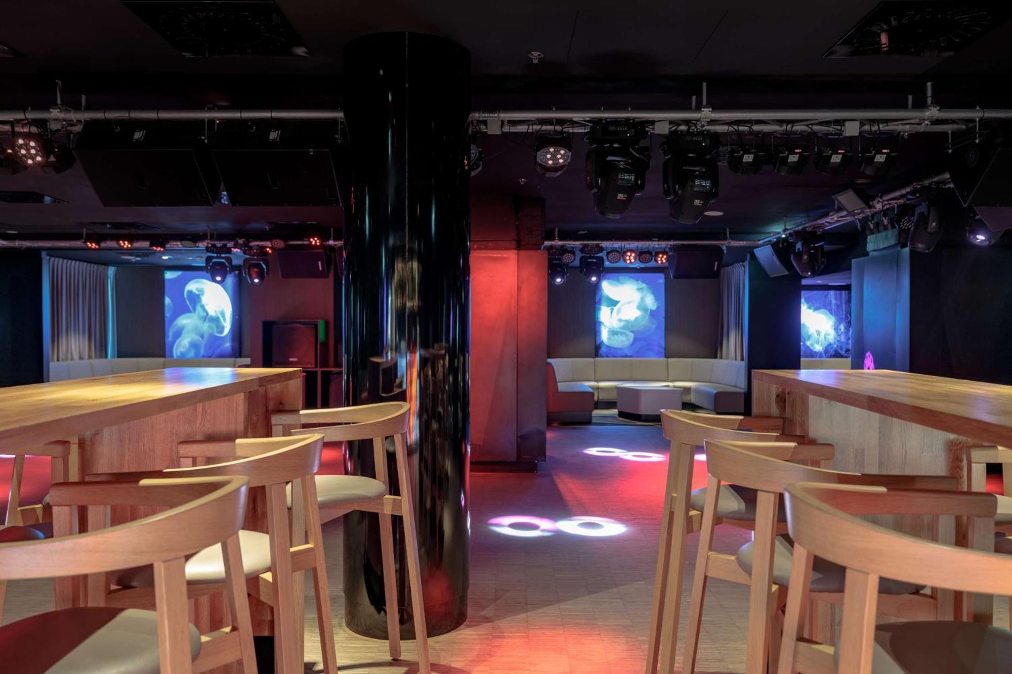 Radisson Blu Resort Świnoujście מראה חיצוני תמונה The photo shows an interior of a modern venue, likely a bar or club. The space features a central black column and light-colored wooden bar stools arranged around a bar area. On the walls, there are large screens displaying vibrant visuals, possibly 