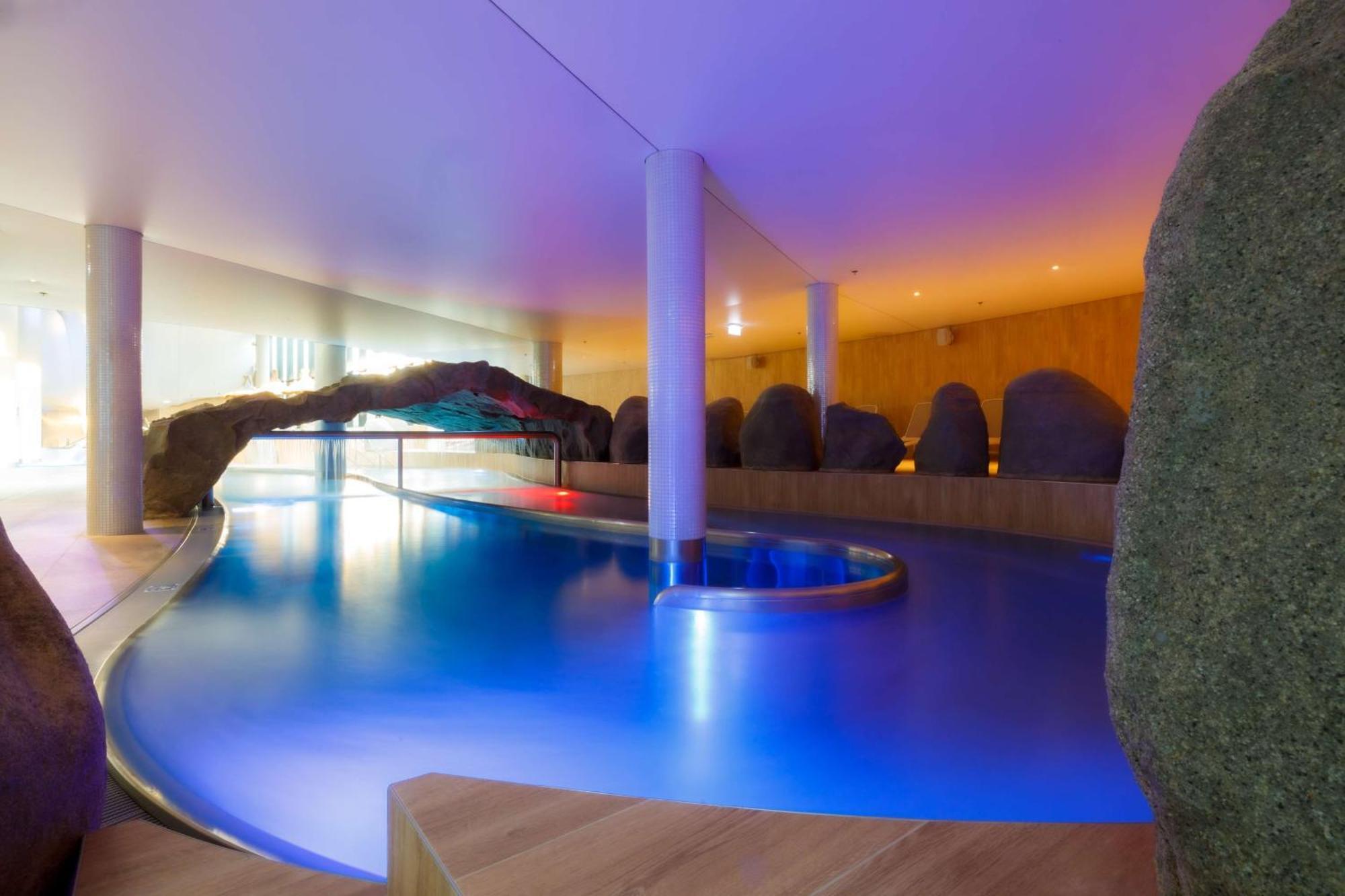 Radisson Blu Resort Świnoujście מראה חיצוני תמונה The photo showcases an elegant indoor swimming pool area. The pool has a flowing, organic shape and is illuminated with soft blue and purple lighting, creating a calming ambiance. Surrounding the pool are smooth, natural-looking stones and wooden ele