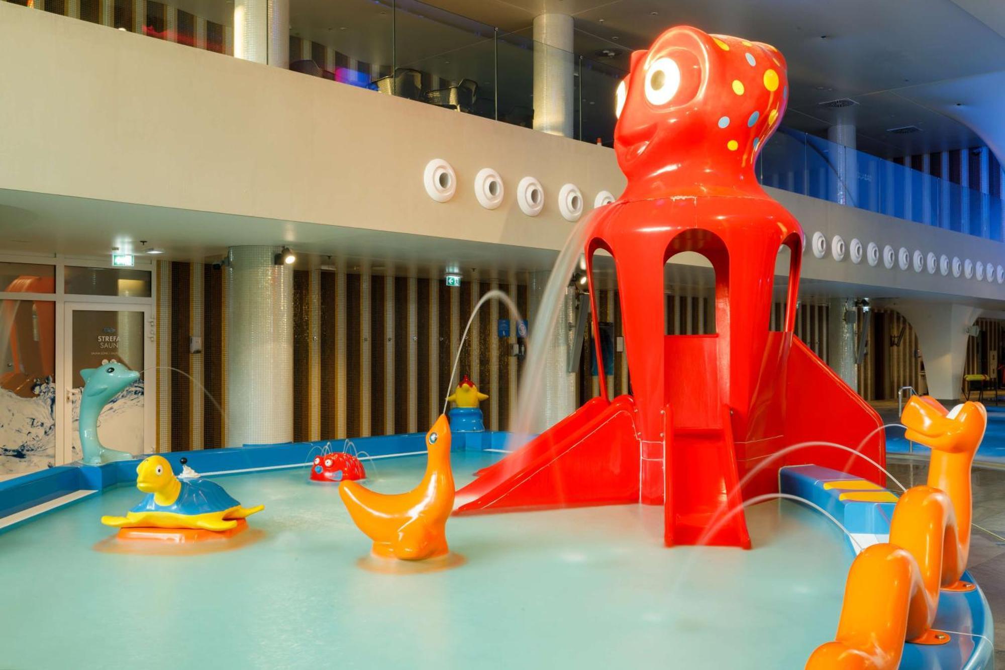Radisson Blu Resort Świnoujście מראה חיצוני תמונה The photo shows a colorful indoor splash pad or water play area designed for children. Central to the scene is a large, bright red octopus structure, which has water fountains spraying out. Surrounding it are various playful, colorful water animals, 